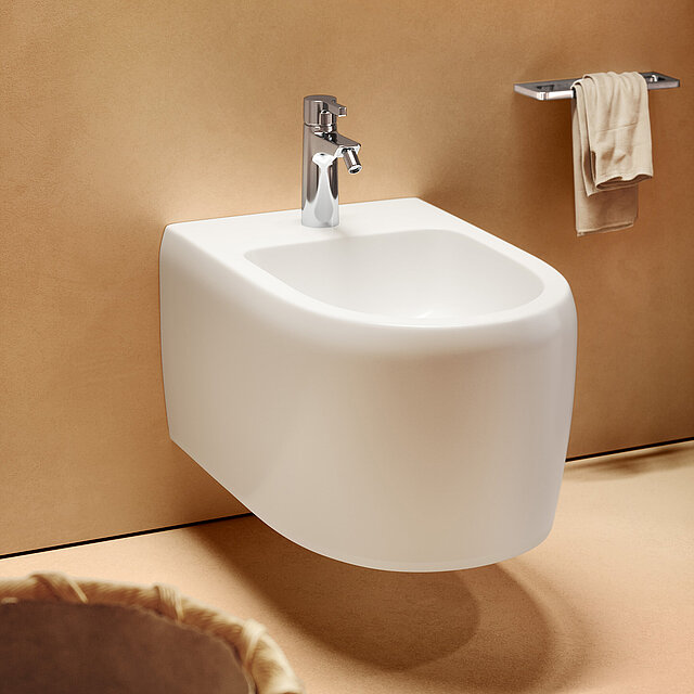 Ein modernes weißes Wandbidet mit einem verchromten Wasserhahn vor einer beigen Wand.