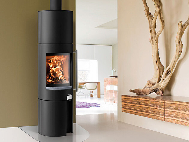 Ein moderner schwarzer Kaminofen mit einer Glastür, durch die man die Flammen im Inneren sehen kann, in einem minimalistischen Wohnraum.