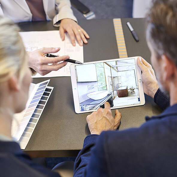 Zwei Personen sitzen an einem Tisch und besprechen einen Einrichtungsplan. Die eine Person hält ein Tablet in der Hand, auf dem ein Badezimmerentwurf abgebildet ist.