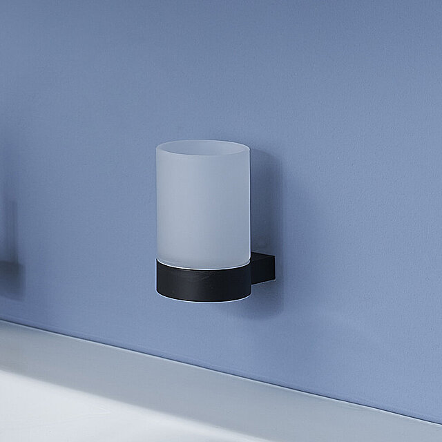 [Translate to Italienisch:] Ein Becher aus Milchglas mit schwarzem Halter ist an einer hellblauen Wand über einer weißen Arbeitsplatte angebracht.