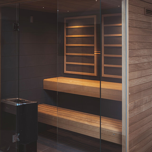 Eine Infrarotkabine mit warmer Beleuchtung und mit zweistufigen Bänken, dunklen Wänden und einer Glastür. 