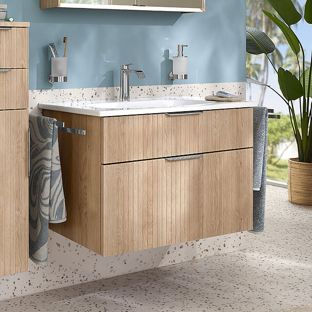 Modernes Badezimmer mit schwebenden Schränken aus hellem Holz und einer weißen Arbeitsplatte vor einer blauen Akzentwand. 