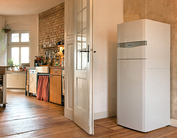 Ein weißes Gas-Brennwert-Kompaktgerät mit integriertem Warmwasserspeicher steht vor einer Wand vor einer Küche mit Holzböden.