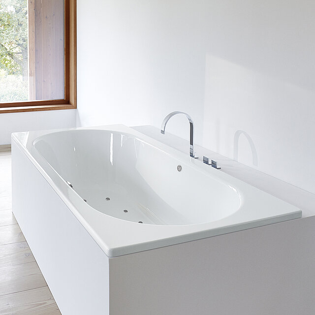 Eine moderne weiße Einbaubadewanne mit Chromarmatur steht in einem minimalistischen Badezimmer.