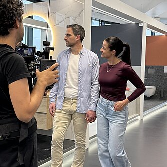 Ein Mann und eine Frau gehen Händchen haltend und lächelnd durch einen modernen Ausstellungsraum, während ein Kameramann sie filmt.