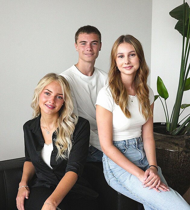 Drei Auszubildende sitzen lächelnd gemeinsam auf einem schwarzen Sofa in einem modernen Raum