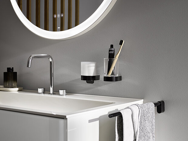 Waschbecken mit einem verchromten Wasserhahn, ein großer runder beleuchteter Spiegel, ein Glas mit Zahnbürste und Zahnpasta und ein Handtuch an einer schwarzen Stange.