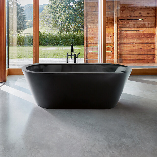 Eine moderne schwarze, freistehende Badewanne steht vor großen Fenstern mit Blick auf grünes Gras, Bäume.