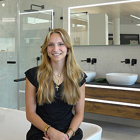 Eine junge Frau mit langen blonden Haaren und schwarzem Outfit sitzt auf dem Rand einer weißen Badewanne in einem modernen Ausstellungsraum.