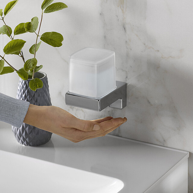 [Translate to Italienisch:] Eine Hand greift unter einen, an der Wand montierten Seifenspender mit gefrostetem Behälter.