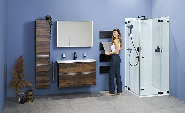 Eine Frau steht in einem Badezimmer mit blauen Wänden, Holzschränken, einem großen Spiegel, einem weißen Waschbecken und einer Glasduschkabine.