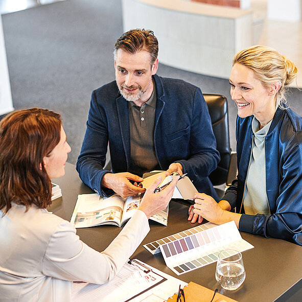 Drei Personen sitzen an einem Tisch und besprechen Dokumente und Farbmuster. 