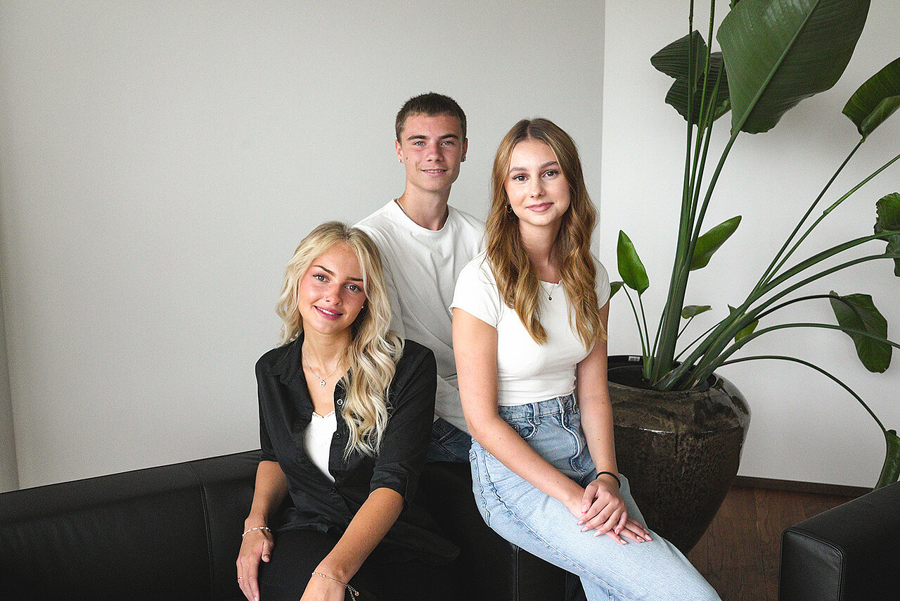 Drei junge Erwachsene sitzen zusammen auf einem schwarzen Sofa in einem modernen Raum mit einer großen Pflanze.