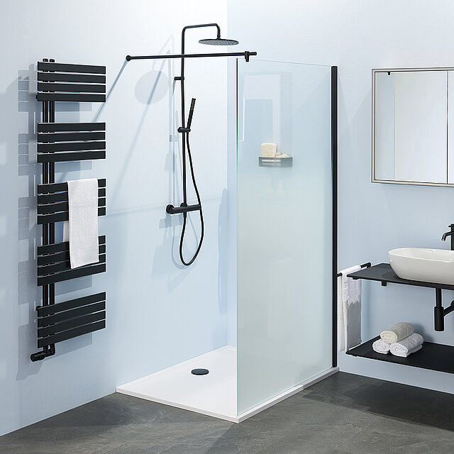 Modernes Bade mit einer begehbaren Dusche mit schwarzem Regenbrausekopf, Trennwand aus Milchglas, schwarzem Handtuchheizkörper mit weißem Handtuch.