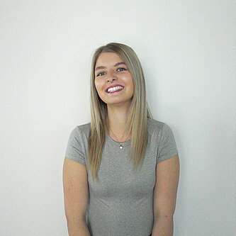 Eine junge Frau mit glattem blondem Haar, die ein graues kurzärmeliges Hemd trägt, steht lächelnd vor einem schlichten weißen Hintergrund. 