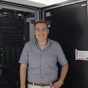 Eine Person mit Brille und kariertem Hemd steht lächelnd vor einem offenen Serverschrank.