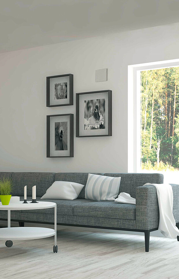 Wohnzimmer mit einem grauen Sofa. Davor steht ein runder Couchtisch mit Kerzen und einer kleinen Pflanze. Ein großes Fenster zeigt die Bäume draußen. 