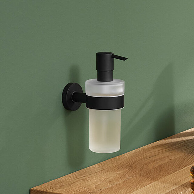 [Translate to Italienisch:] Ein wandmontierter Seifenspender aus Milchglas mit schwarzer Pumpe und Halterung ist an einer Wand angebracht.