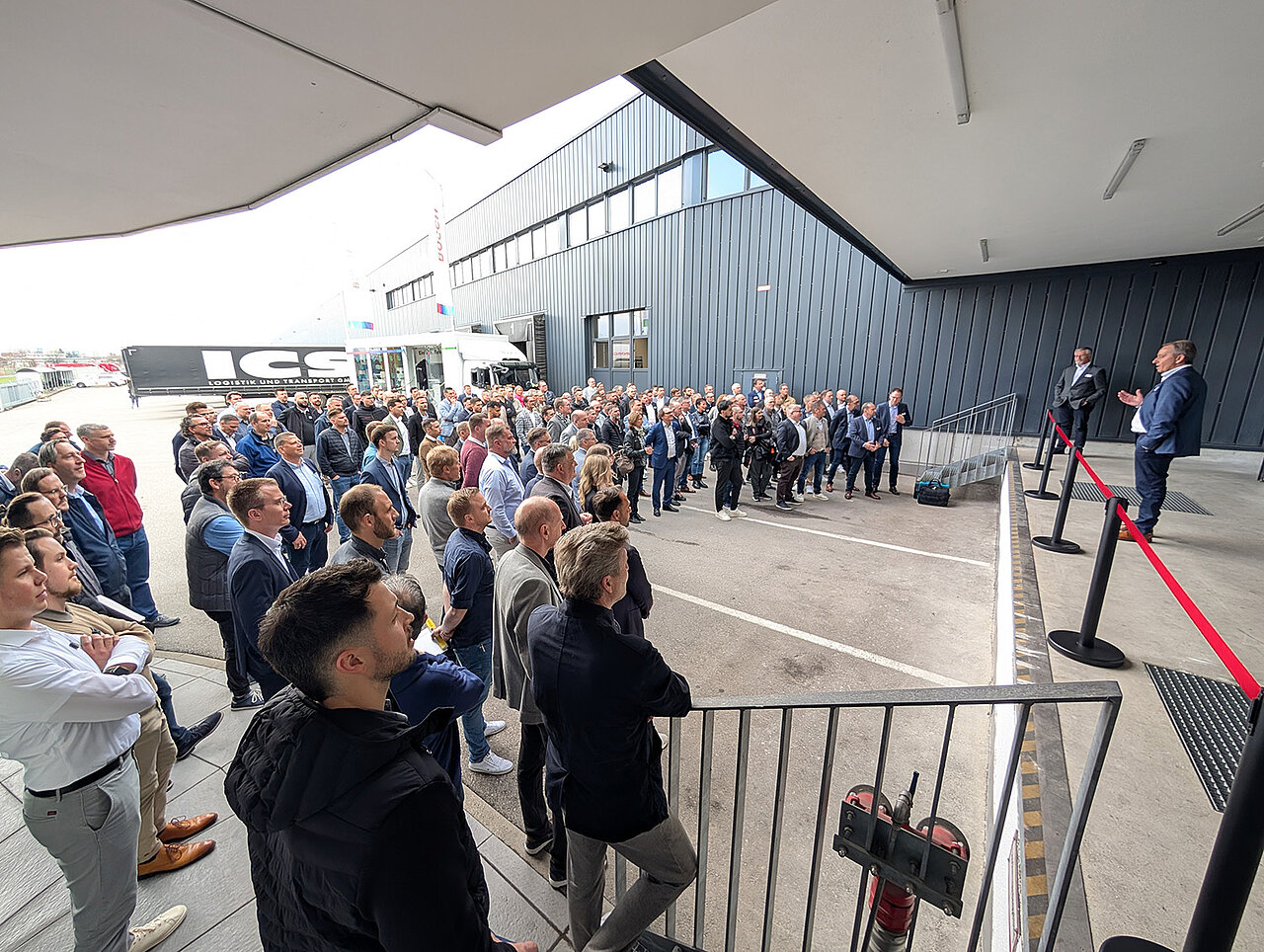 Eine große Gruppe von Menschen steht vor einem modernen Gebäude und hört aufmerksam einem Redner zu.