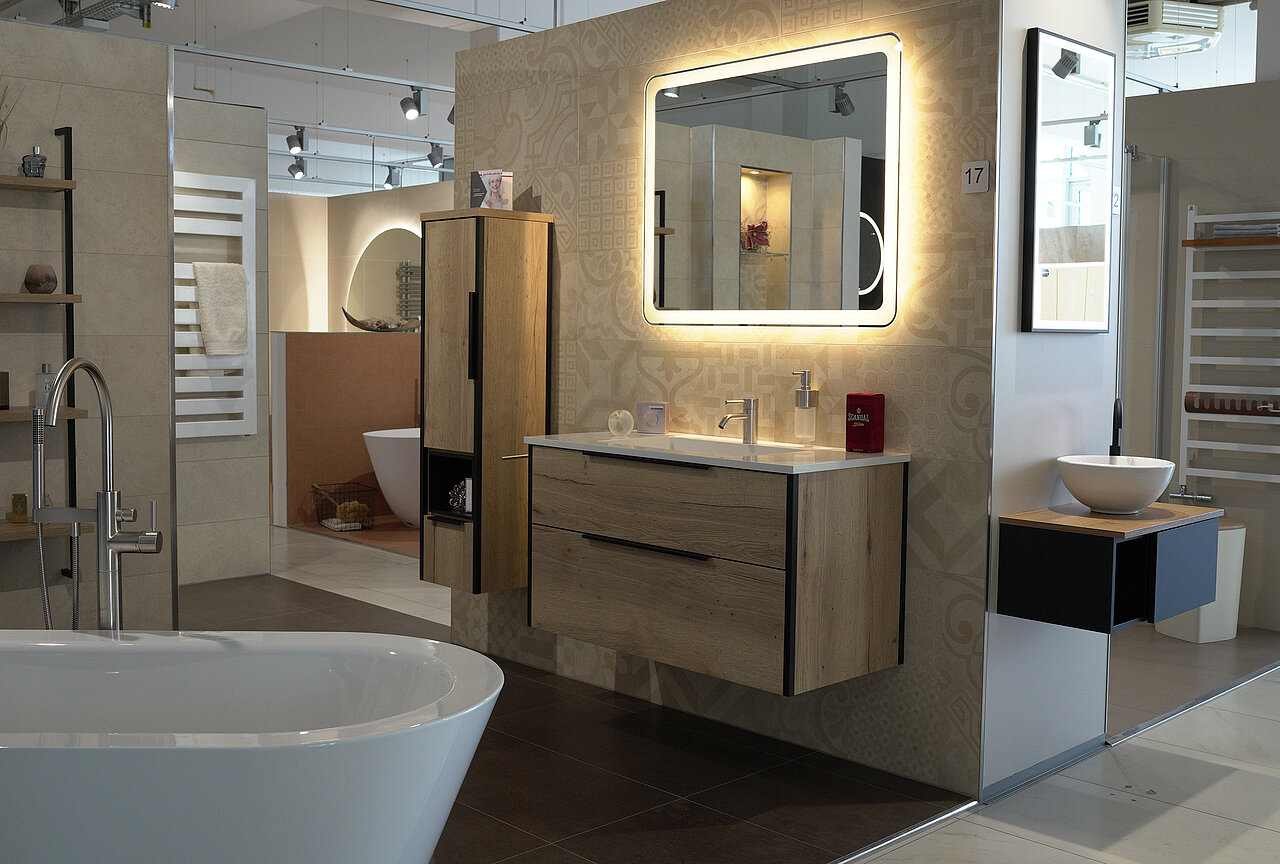 Badezimmerausstellung mit Waschtisch und Waschbecken, einem großen beleuchteten Spiegel, einem vertikalen Schrank und einer weißen freistehenden Badewanne.