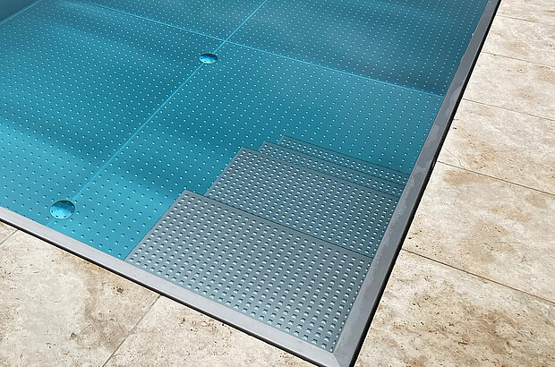 Ein Edelstahlpool mit hellblauem Wasser. Versenkte Stufen mit perforierter, rutschfester Oberfläche führen ins Becken. Hellbraune Steinfliesen umrahmen das Schwimmbecken.
