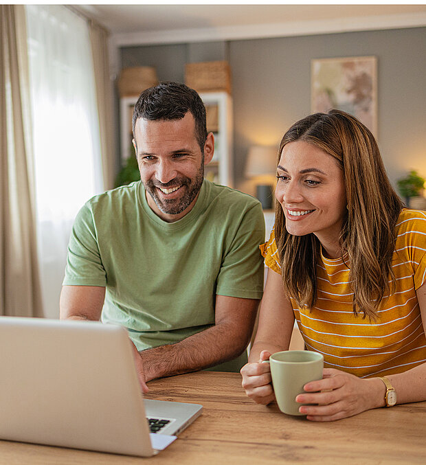 Ein lächelnder Mann und eine lächelnde Frau sitzen an einem Tisch und schauen auf einen Laptop-Bildschirm. 
