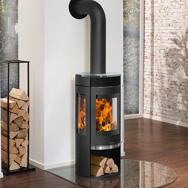 Schwarzer Holzofen mit Glastür und sichtbaren Flammen auf Glasbodenplatte, daneben Brennholzstapel in hellem Raum.