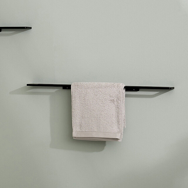 [Translate to Italienisch:] Ein minimalistischer schwarzer Handtuchhalter an einer hellgrauen Wand, der ein ordentlich gefaltetes hellgraues Handtuch hält.
