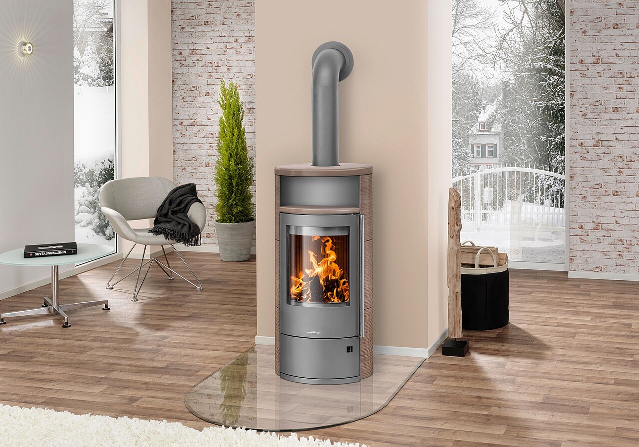 Modernes Wohnzimmer mit Holzofen auf Glas-Funkenschutz, Holzboden, weißem Sessel, Pflanze und Blick ins verschneite Freie.