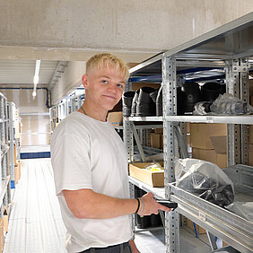 Ein junger Mann mit blondem Haar und weißem T-Shirt steht in einem Lagerraum, lächelt in die Kamera 
