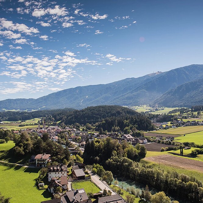Ansicht von St. Lorenzen: Ein kleines Dorf mit Häusern und Feldern liegt eingebettet in ein grünes Tal, umgeben von bewaldeten Hügeln und Bergen unter blauem Himmel.