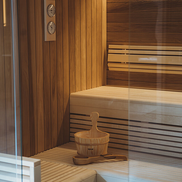 Eine Holzsauna mit einer hellen Holzbank, passenden Wänden, einem Eimer und einer Schöpfkelle auf der Bank.