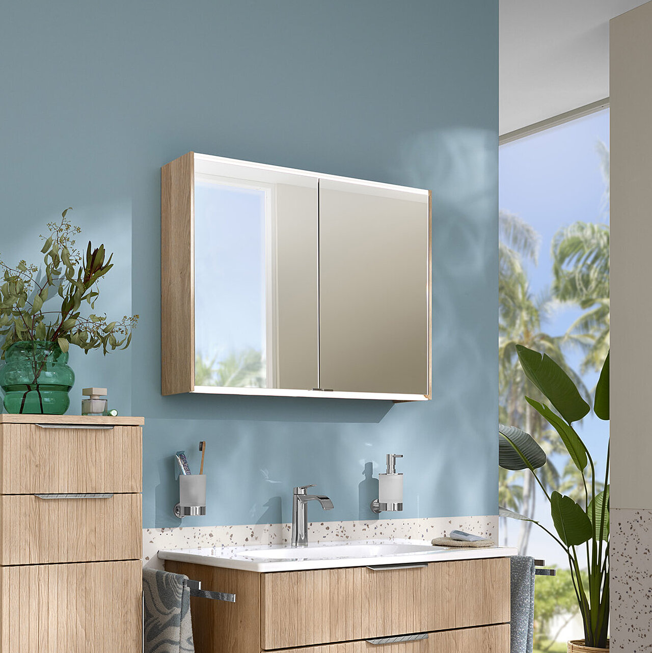 Modernes Bad mit schwebenden Schränken aus hellem Holz, blauer Akzentwand, Terrazzoboden und einem großen Spiegel.